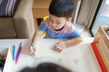 Asyalı genç bir çocuk, annesiyle birlikte renkli bir kalemle bir kağıt çiziyor..