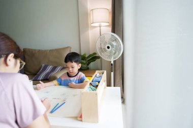 Asyalı anne renkli kalemle çizim yapan bir çocuğa yardım ediyor. Çocuk sarı bir sandalye ve beyaz bir masa kullanıyor.