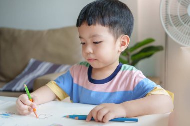 Asyalı çocuk bir masada oturuyor ve kağıt üzerinde renkli kalemle boyuyor. Yaratıcılık ve oyunculuk kavramı