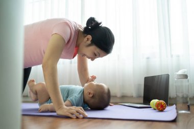 Asyalı anne oturma odasında yoga minderinde bebekle oynuyor, neşeli bir egzersiz yapıyor, neşeli bir bağ kuruyor.