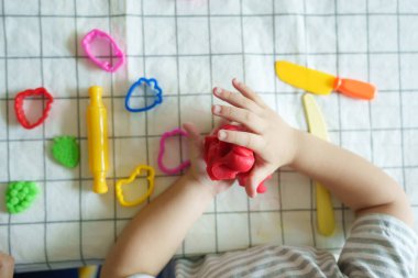 Üst görünüm, renkli plastik kalıplar ve araçlarla çevrili kareli bir masa örtüsü üzerinde kırmızı modelleme kilini yoğuran bir çocuğun yakın çekimi