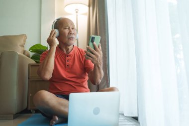 Asyalı yaşlı bir adam dizüstü bilgisayarının önünde oturuyor, kulaklık takıyor ve birlikte şarkı söylerken akıllı bir telefon tutuyor..