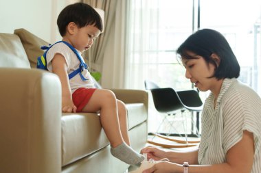 Annesi ayakkabılarını giymesine yardım ederken Asyalı genç çocuk sırt çantasıyla kanepede oturuyor. Okula hazırım..