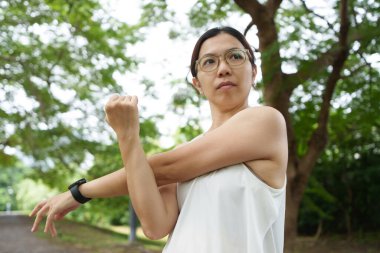 Asyalı kadın parkta yapılan bir egzersiz sırasında kollarını göğsüne uzatıyor. Odaklanmış ifade, aktif yaşam tarzı ve egzersiz rutini.