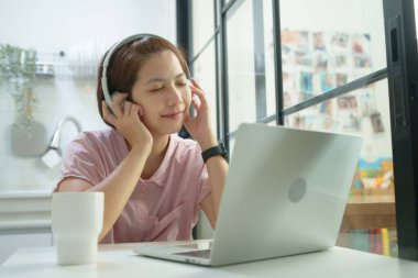 Asyalı genç kadın masasında oturan kulaklıklarla ve laptopuyla rahatlatıcı müzik dinliyor..