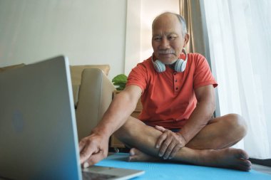 Kulaklık takan Asyalı yaşlı adam laptopunun önünde yoga minderinin üzerinde oturuyor..
