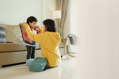 Asyalı bir anne, küçük oğluna oturma odasında süveter giymesinde yardım ediyor. Okul konseptine dönelim..