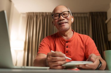 Evde tablet kullanırken gülümseyen Asyalı yaşlı adamın portresi, gözlük ve kablosuz kulaklık takıyor..