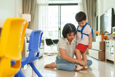 Asyalı anne, genç oğlunun parlak bir oturma odasında ayakkabı giymesine yardım ediyor. Okula hazırlanmak.