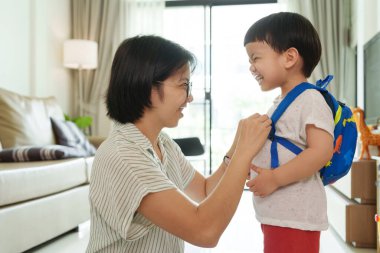 Asyalı anne, küçük oğlunun sırt çantalarını oturma odasında gülümserken düzeltir.