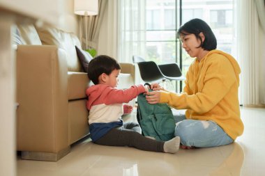 Asyalı anne, oturma odasında yerde otururken küçük oğlunun yeşil sırt çantasını toplamasına yardım ediyor. Açık hava aktivitesi ya da okul için hazırlanın..