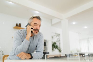 Orta yaşlı bir adam, rahat bir ifadeyle akıllı bir telefonla konuşuyor, modern bir mutfağın içinde mutfak tezgahına yaslanıyor..