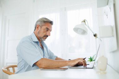 Olgun bir adam, beyaz bir odadaki masanın üstünde lambayla ev ofisinin bilgisayarında çalışıyor..