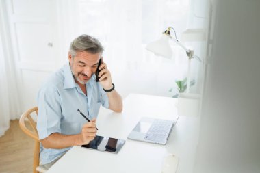 Son sınıf öğrencisi akıllı telefondan konuşuyor, tablet kullanıyor, gülümsüyor ve parlak bir ev ofisinde çalışıyor..