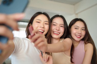 Üç genç Asyalı kadın akıllı telefonlarıyla neşeli bir selfie çekiyor. Güzel kızlar gülümsüyor ve yakından poz veriyor, bir anlık dostluk ve neşe yakalıyorlar..