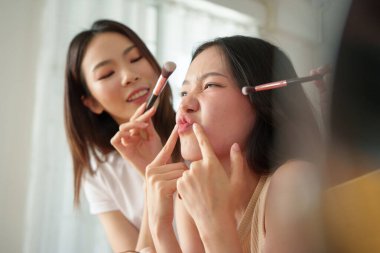 Makyaj yapan iki genç Asyalı kadın gülümseyen ve komik suratlar yapan ergen kız bir arkadaşlık ve güzellik anı yakalıyorlar..