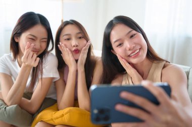 Üç genç Asyalı kadın neşeli bir selfie çekiyorlar anlamlı yüzlerle poz veriyorlar, dostça gülümseme ve el hareketleri, oturma odasında akıllı telefonla eğlenceli bir bağ kurma anı yakalıyorlar..