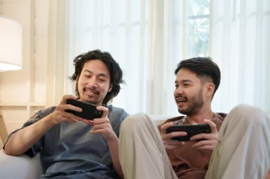 İki Asyalı adam kanepede oyun oynuyor. İki arkadaş gülümsüyor ve akıllı telefondan dostça bir yarışmanın tadını çıkarıyor..