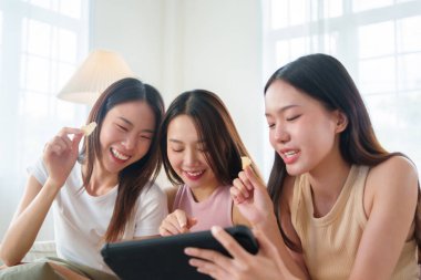 Üç genç Asyalı kadın gülümseyip abur cubur yerken dijital tabletin içeriğiyle uğraşıyorlar. Güzel kızlar birlikte rahat ve neşeli bir anın tadını çıkarıyorlar..