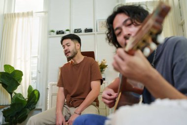 Genç adam gitar çalarken, arkadaşı rahat, kapalı bir yaşam alanında şarkı söylüyor, rahat ve samimi bir müzik anı yakalıyor..