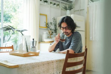Gözlüklü ve sakallı genç Asyalı bir adam masada kahve kokluyor ve içiyor. Japon erkek rahat bir yemek masasında bir fincan kahveyle sakin bir anın tadını çıkarıyor.