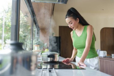 Genç Asyalı kadın modern bir mutfakta yemek pişiriyor. Buhar yükselirken tencereyi dikkatle izliyor. Etrafı sakin ve rahat bir ortamla çevrili..