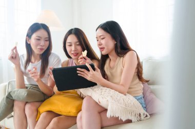 Üç genç Asyalı kadın oturma odasındaki bir kanepede dijital tablette bir şey izlerken abur cubur yiyor. Arkadaşlık kavramı. Güzel Koreli, Japon ve Çinli kızlar.