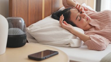 Genç Asyalı kadın, gözleri kapalı ve yorgun bir şekilde yatakta uzanıyor ve elini başının üstünde dinlendiriyor, akıllı telefonu da komodinin yanında..