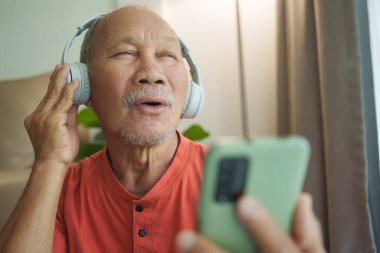 Turuncu ya da kırmızı tişörtlü mutlu Asyalı adam kablosuz kulaklıkla müzik dinliyor ve akıllı telefon ya da cep telefonuyla birlikte şarkı söylüyor..