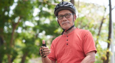 Turuncu tişörtlü Asyalı yaşlı bisikletçi, kask ve eldiven giyerken başparmağını kaldırıyor, yeşil bir parkta güvenle dikiliyor, aktif bir yaşam tarzının tadını çıkarıyor.