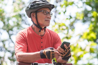 Parkta bisikletle bisiklet sürerken kasklı, cep telefonu ya da akıllı telefon taşıyan Asya 'lı yaşlı Çinli bir adamın portresi. Büyükbabam bisiklete binerek egzersiz yapıyor. Uzaklara bakıyorum..