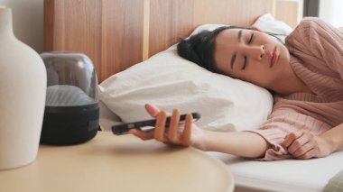 Yatakta uzanan Asyalı kadın yatak odasında rahat pembe bir kazak giyerek komodindeki akıllı telefona uzanıyor.