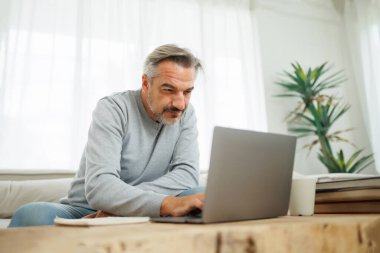 Sakallı, olgun bir adam. Parlak bir oturma odasında dizüstü bilgisayarla çalışıyor..