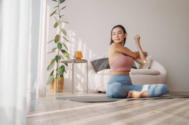 Huzurlu bir ev ortamında uzanan genç Asyalı kadın, doğal ışık ile oturma yoga duruşu sergiliyor, sakin ortamı geliştiriyor.