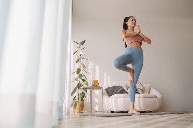 İnce Asyalı kadın, ağaç yoga pozu veriyor, dengeyi ve minimalist bir odada duruşu temsil ediyor.