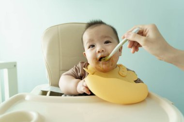 Asyalı bebek kaşıkla yüksek sandalyede besleniyor, püre yiyor, yemek yiyor.