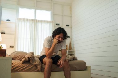 Genç Asyalı bir adam düzgün yapılmış bir yatağın kenarında oturuyor, yüzü derin düşüncelere dalmış bir ifadeyle dinleniyor, parlak ve minimalist bir yatak odasıyla çevrili..