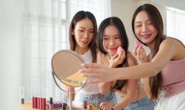Üç Asyalı kadın güzellik süngeri kullanıyor ve evde cilt bakımı yapıyor. Tahta bir masadaki kozmetik eşyalarla çevrili aynaya bakıyorlar..
