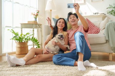 Gülümseyen Asyalı çift, köpekleri ve akıllı telefonlarıyla oturma odasında selfie çekiyor.