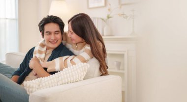 Asyalı bir çift kanepede oturuyor, şefkatle gülümsüyor, yumuşak ışıklandırmalı ve zarif dekorlu rahat bir oturma odasında kucaklaşıyorlar..