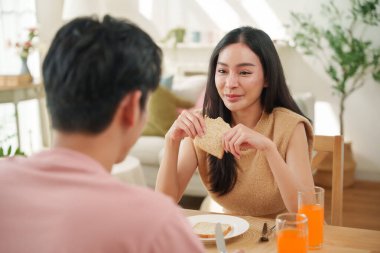 Parlak doğal ışık ve Sevgililer Günü havasıyla çevrili sıcak bir kahvaltı sırasında yemek masasında erkek arkadaşıyla sohbet ederken gülümseyen Asyalı kadın..