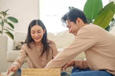 Genç Asyalı çift ellerinde açık bir sepetle yerde oturmuş sıcak dekore edilmiş bir oturma odasında yaratıcı bir aktivite düzenliyor ve eğleniyorlar. Taşınma konsepti