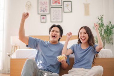 Neşeli genç Asyalı çift oturma odalarının zemininde oturmuş, spordan zevk alırken ve patlamış mısır paylaşırken tezahürat yapıyor ve gülümsüyorlar..