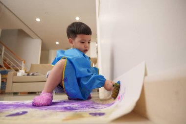 Mavi önlüklü Asyalı çocuk ev ortamında canlı renkler kullanarak yerdeki büyük kağıt parçalarına resim yapıyor. Çocuk odaklanmış ve yaratıcı faaliyetlerle meşgul.