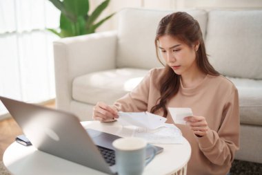 Oturma odasındaki kahve masasında oturan Asyalı kadın, bir evde dizüstü bilgisayarla çevrili finansal belgeleri dikkatle inceliyor..