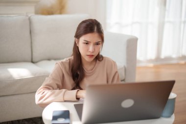 Ciddi ve odaklanmış bir ifadeyle evden çalışan Asyalı bir kadın, oturma odasında bir kahve masasında dizüstü bilgisayarını kullanarak oturuyor..