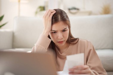 Asyalı kadın mali belgeleri incelerken stresli hissediyor, oturma odasındaki masada hayal kırıklığı içinde başını tutuyor..