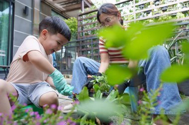 Asyalı anne ve küçük oğlu, yemyeşil ve renkli çiçeklerle çevrili güneşli bir günde neşeyle bahçeyle uğraşıyorlar. Çocuk eldiven takıyor ve bitkilere yardım ediyor.