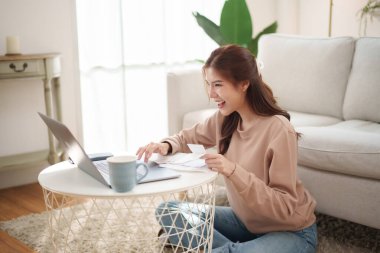 Evde finansman yönetirken dizüstü bilgisayar kullanan gülümseyen genç Asyalı kadın, oturma odasında yerde oturuyor, elinde yuvarlak bir masanın fişi var.