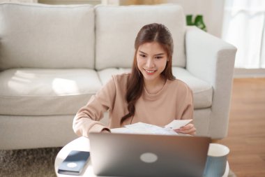 Evde bilgisayarla çalışırken gülümseyen Asyalı bir kadın. Etrafı mali belgelerle çevrili.